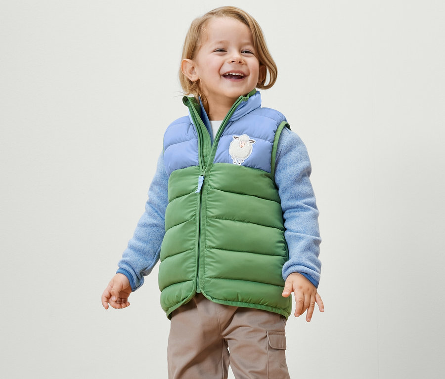 Lächelnder kleiner Junge trägt eine Kinder-Steppweste mit Colorblocking, einen blauen Pullover und eine Hose.