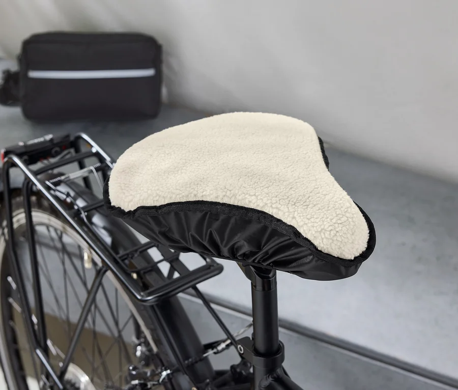 Detailaufnahme eines Fahrrads mit schwarzem Rahmen, dessen Sattel mit einem beigen, fellartigen Bezug versehen ist. Im Hintergrund befindet sich eine schwarze Tasche mit einem weißen Streifen.