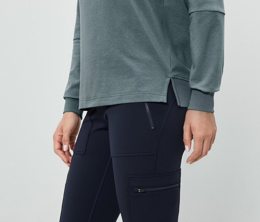 Detailaufnahme einer Frau in marineblauen Hosen mit Reißverschlusstaschen und einem blaugrünen Langarmshirt.