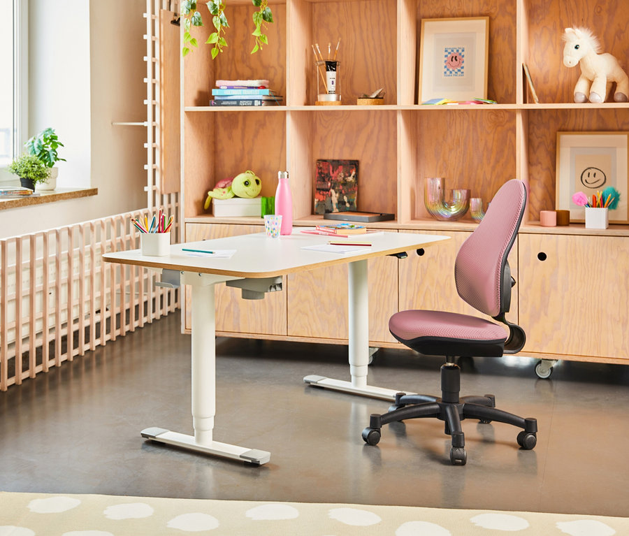 Ein weißer Schreibtisch steht in einem Raum mit einem hölzernen Bücherregal und einem rosa Topstar Stuhl »Open Art Junior«.