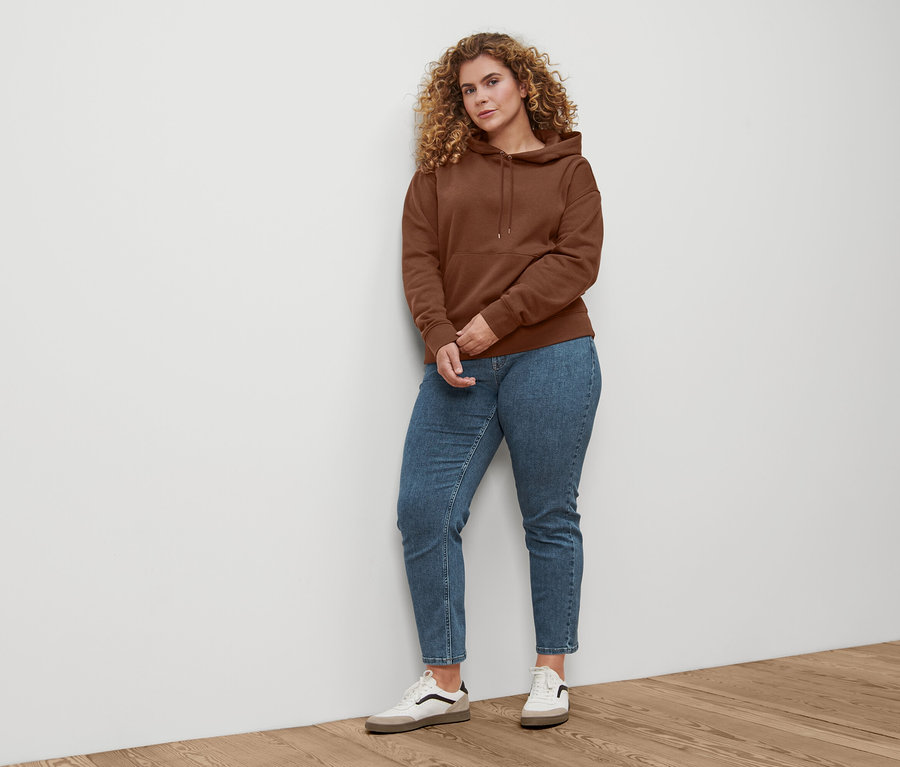 Eine Frau steht und trägt Straight-Denim »Fit Juna« in Midblue, Leder-Sneaker und einen dunkelbraunen Hoodie.