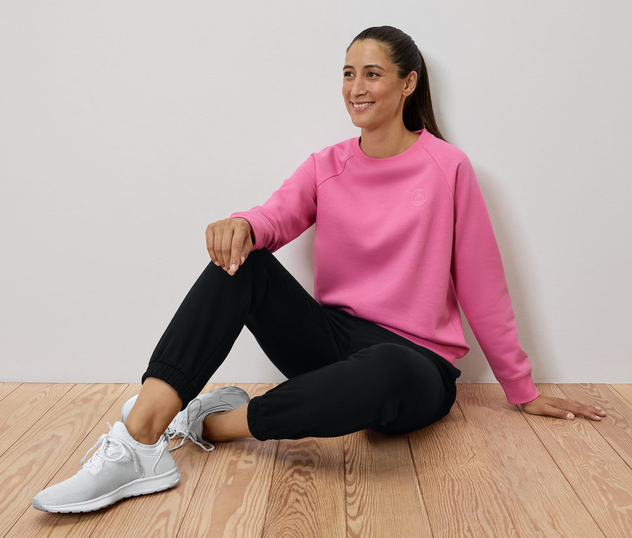 Frau sitzt auf Holzboden, trägt ein rosa Sweatshirt, schwarze Hosen und weiße Turnschuhe.