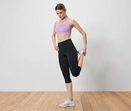 Frau dehnt ihr Bein, indem sie ihren Knöchel in der Hand hält. Sie trägt einen lila Sport-BH, schwarze Leggings und weiße Turnschuhe.
