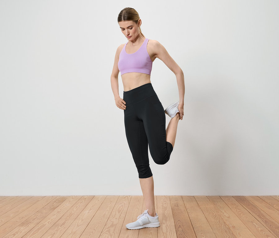 Frau dehnt ihr Bein, indem sie ihren Knöchel in der Hand hält. Sie trägt einen lila Sport-BH, schwarze Leggings und weiße Turnschuhe.