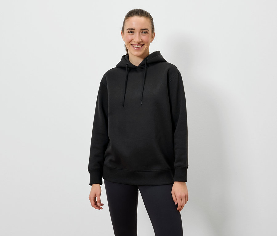 Frau mit schwarzem Hoodie und schwarzen Leggings steht vor einem weißen Hintergrund.
