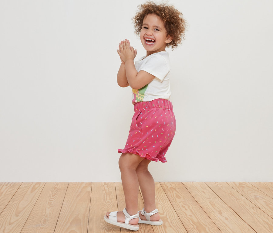 Lächelndes lockiges Mädchen klatscht in die Hände und trägt Kinder-Sweat-Shorts.