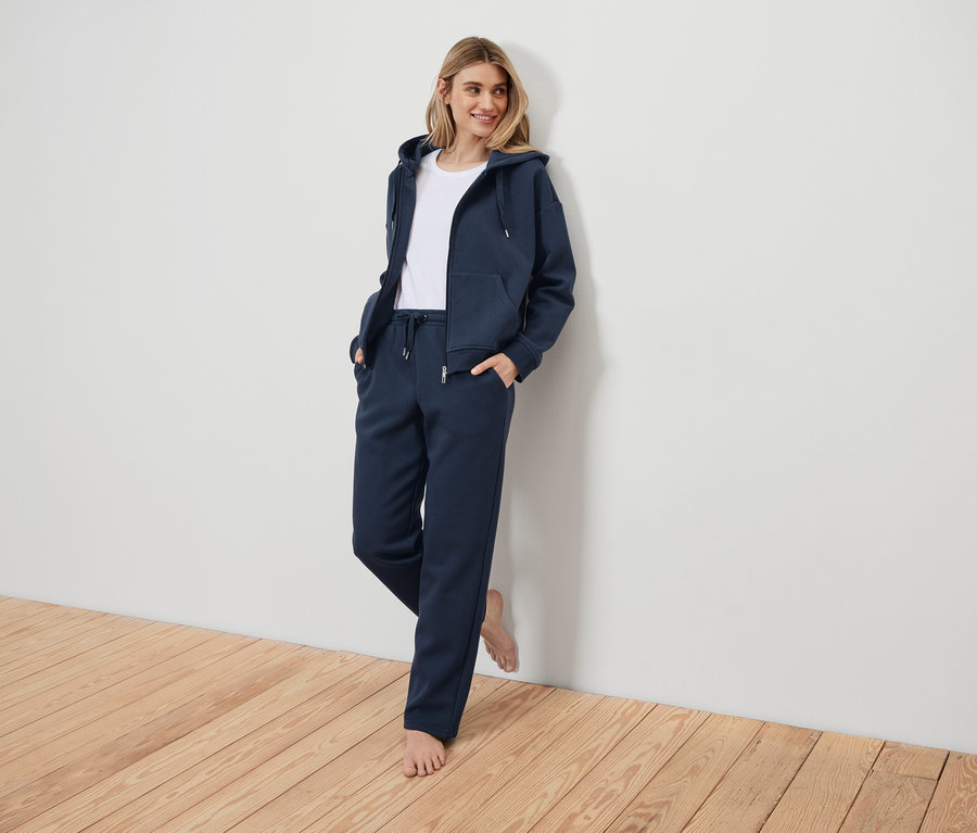 Frau in dunkelblauem Trainingsanzug, an eine weiße Wand gelehnt.