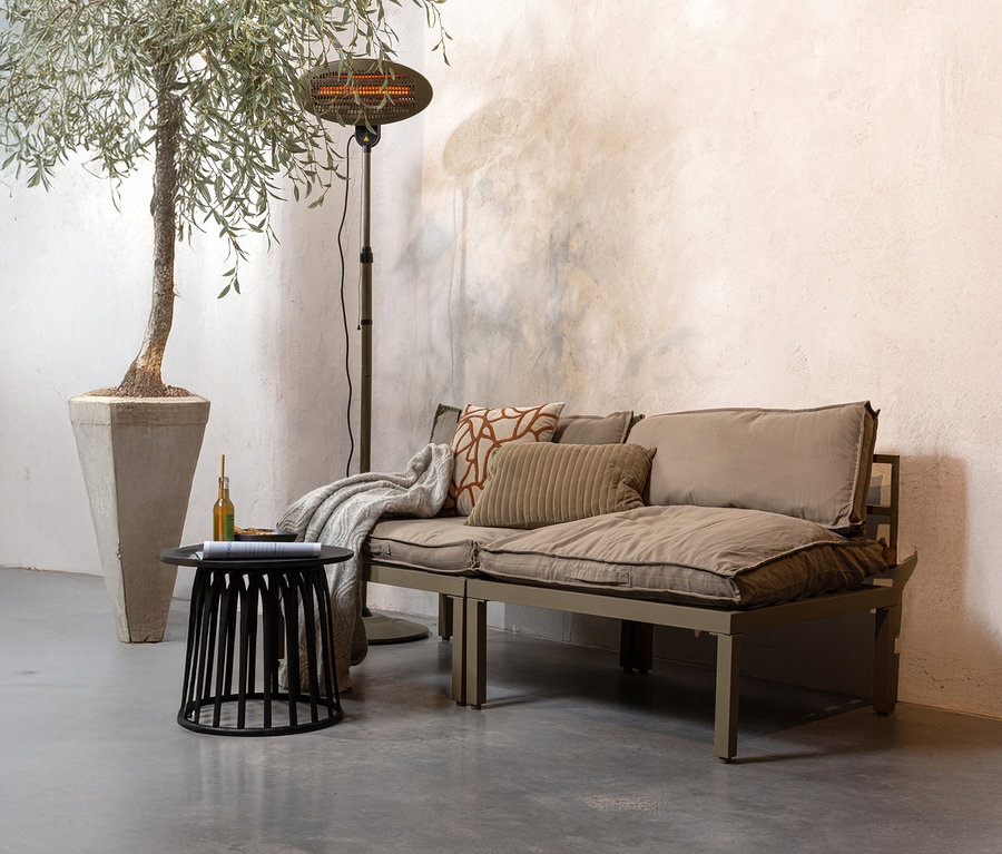 Ein beiges Sofa mit Kissen und einer Decke steht neben einem schwarzen Tisch mit einer Flasche und einem Buch. Dahinter stehen eine Stehlampe und ein Olivenbaum im Topf.