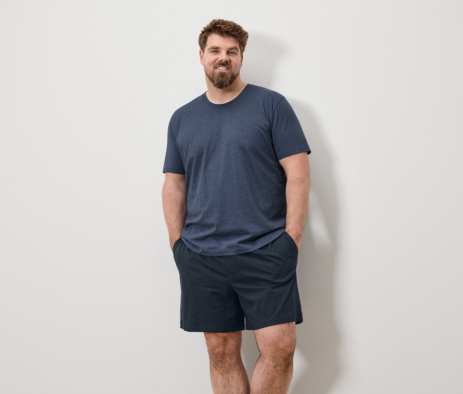 Ein Mann mit Bart steht mit den Händen in den Taschen vor einer weißen Wand, er trägt ein blaues T-Shirt und Shorts.