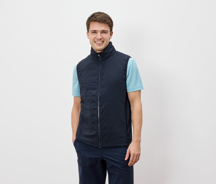 Ein Mann lächelt in die Kamera und trägt eine marineblaue Weste über einem hellblauen T-Shirt.