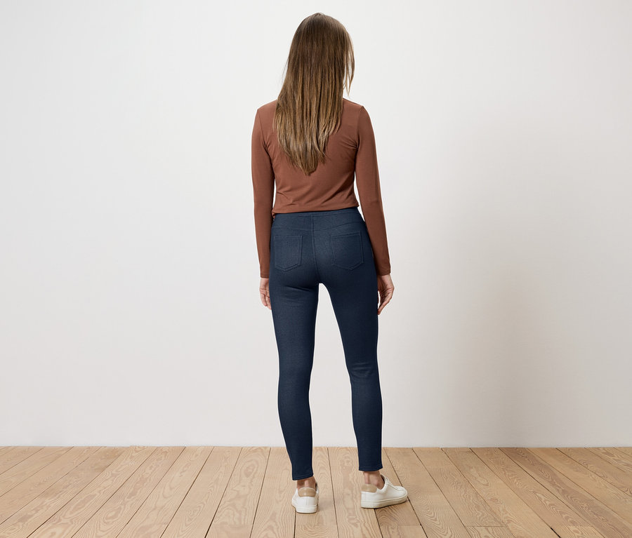 Eine Frau mit braunen Haaren steht mit dem Rücken zum Betrachter, trägt ein braunes Langarmshirt, blaue Jeans und weiße Schuhe.