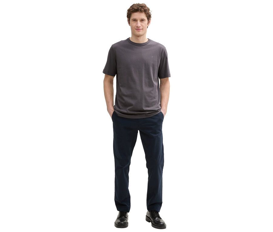Ein Mann mit lockigem Haar steht mit den Händen in den Hosentaschen, er trägt ein graues T-Shirt, blaue Hose und schwarze Schuhe.