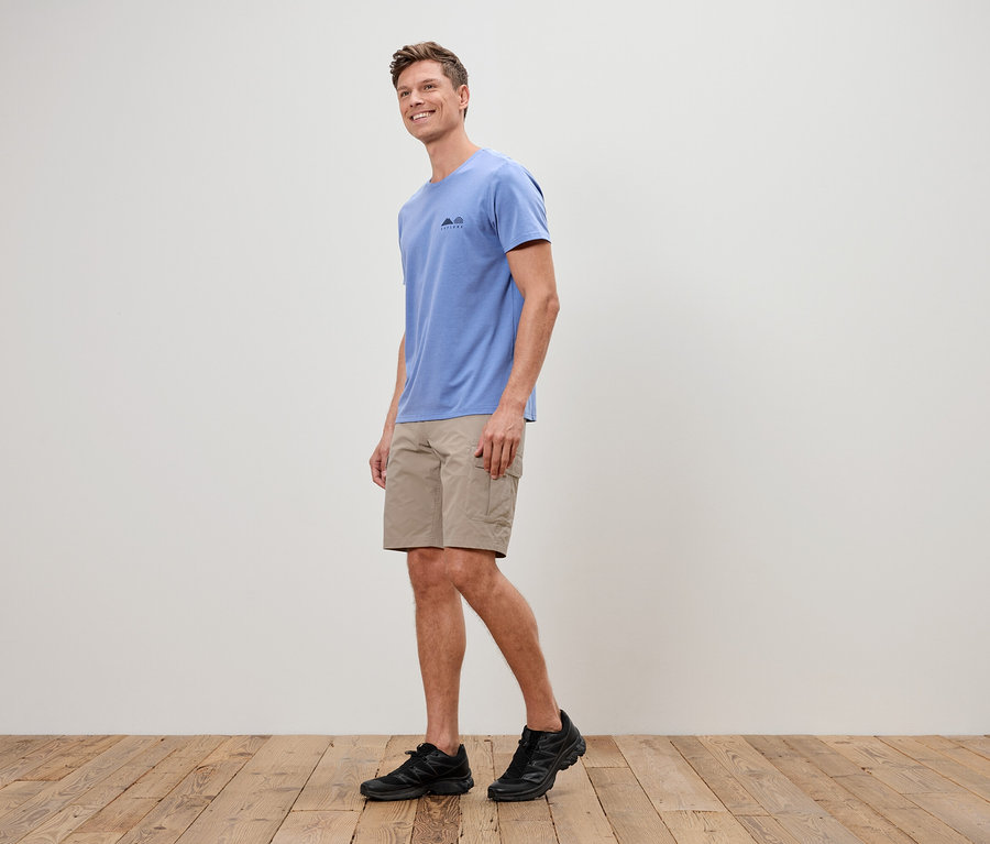 Ein Mann geht auf einem Holzboden und trägt ein blaues T-Shirt, kurze, beigefarbene Hosen und schwarze Turnschuhe.