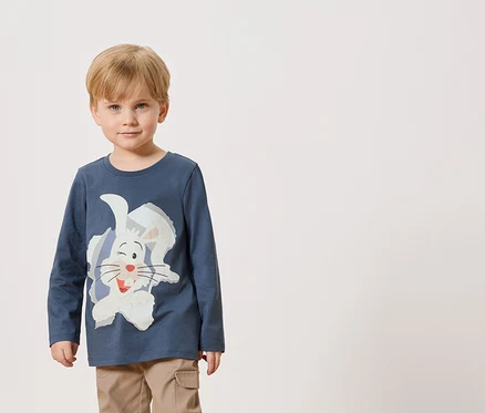 Ein kleiner Junge trägt ein blaues Langarmshirt mit einem weißen Hasenaufdruck und eine beigefarbene Hose.
