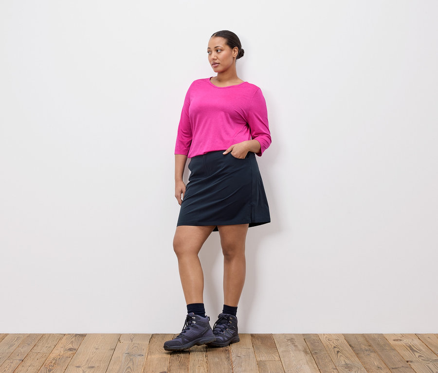 Frau mit pinkem Oberteil, dunklem Rock und Wanderschuhen steht vor einer weißen Wand.