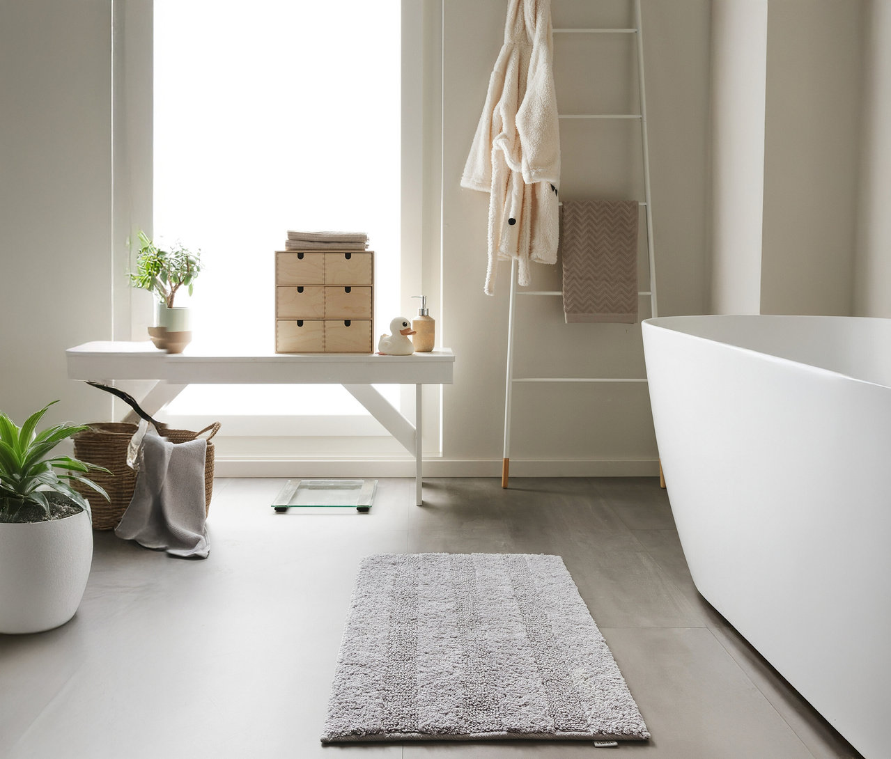 Eine hellgraue benuta Badematte »Ilka«, 50 x 80 cm liegt auf einem grauen Badezimmerboden.