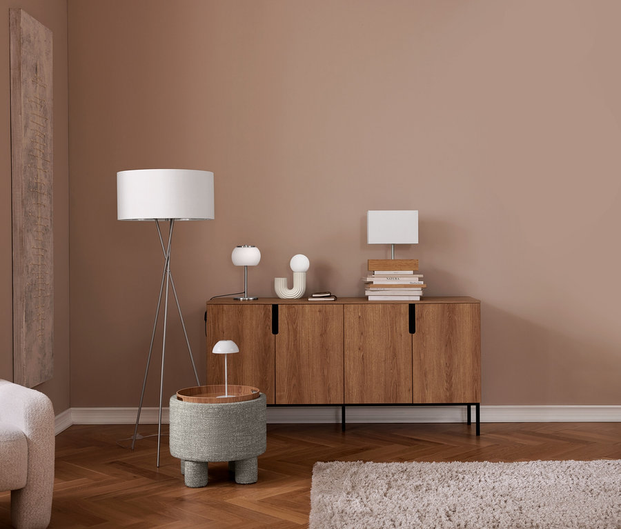 Ein modernes Wohnzimmer mit einem hellbeigen Sessel, einem Fischgrätparkettboden und einer altrosafarbenen Wand. Eine silberne Dreibein-Stehlampe steht neben einem grauen, texturierten Ottoman mit Holztablett und einer kleinen weißen Lampe. Ein Holzsideboard trägt zwei weiße Tischlampen, Deko-Objekte und Bücherstapel. Ein heller, zotteliger Teppich liegt am Boden.