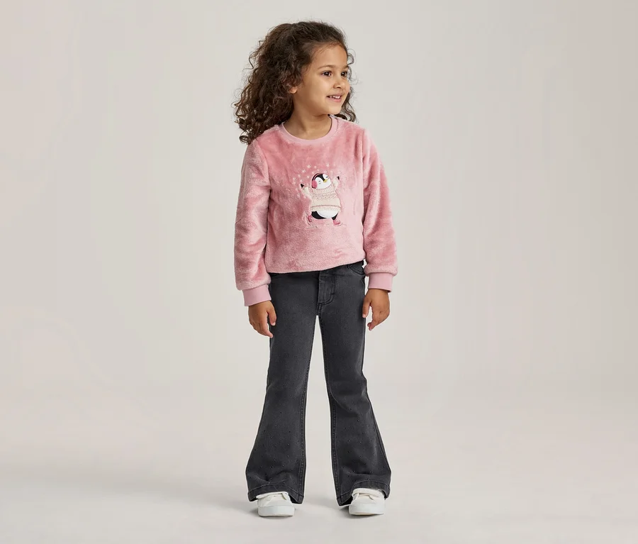 Junges Mädchen mit lockigem Haar steht da und trägt einen rosa flauschigen Pullover mit Pinguin-Motiv und dunkelgraue Schlagjeans.