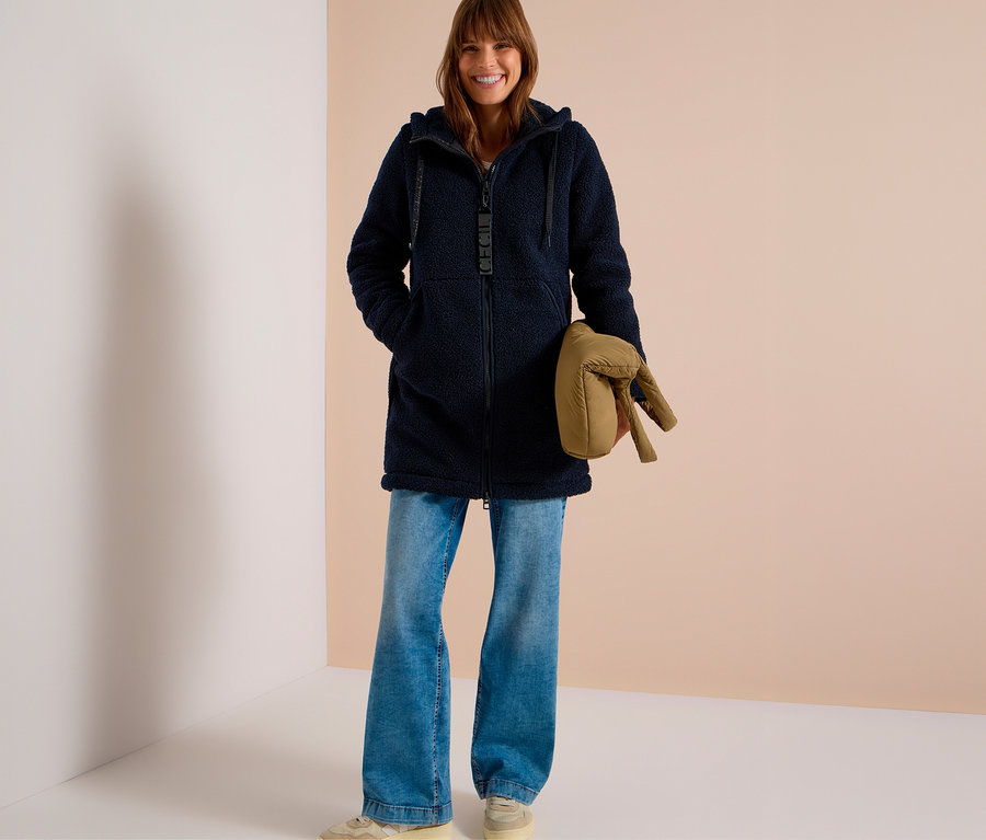 Lächelnde Frau in einer dunkelblauen, flauschigen CECIL Jacke mit Kapuze, hellblauen Jeans und beigen Sneakern, hält eine beige Steppjacke.