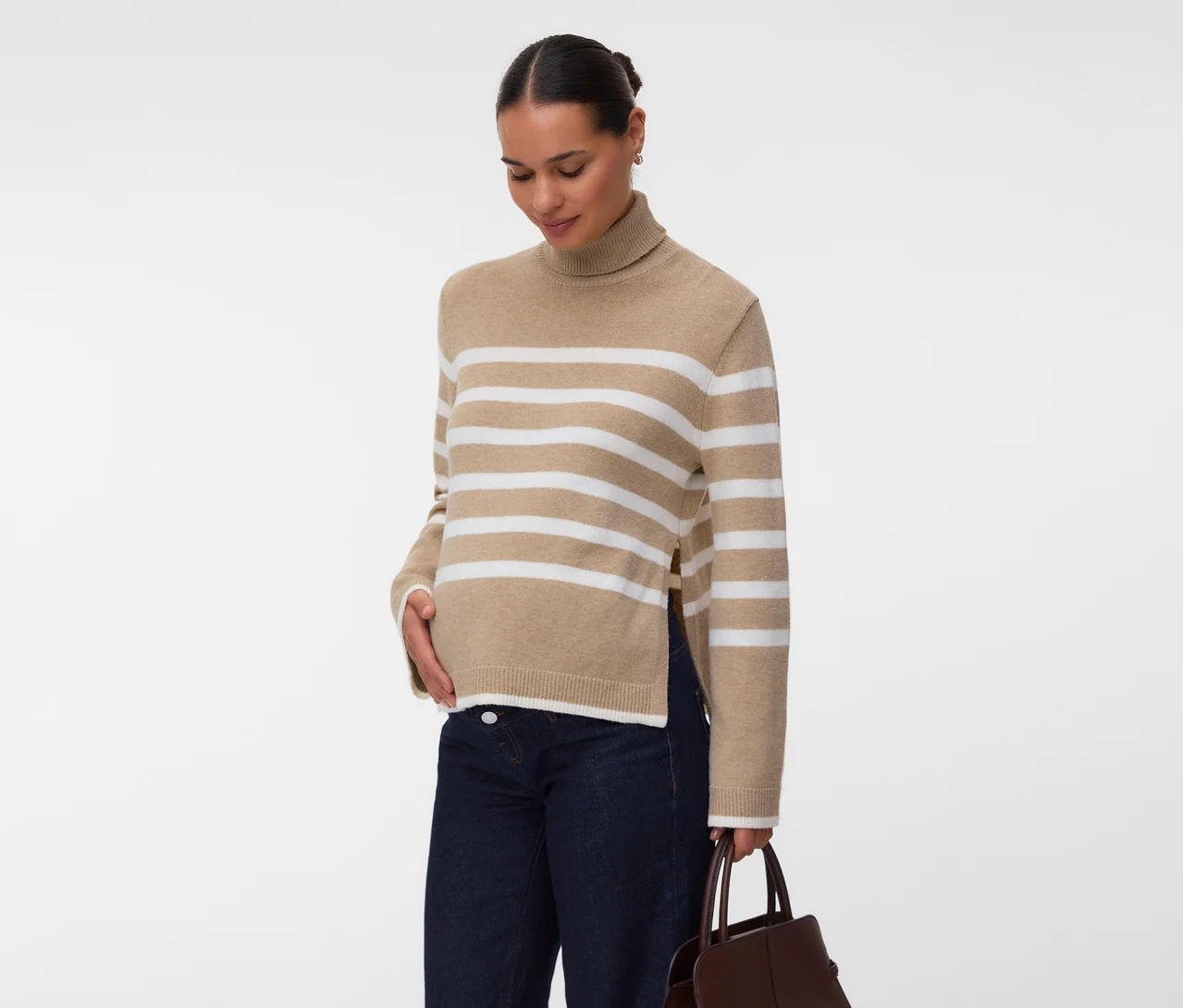 Frau in einem beigefarbenen Rollkragenpullover mit weißen Streifen und Seitenschlitzen, dunklen Jeans und einer braunen Handtasche, die Hand auf dem Bauch.