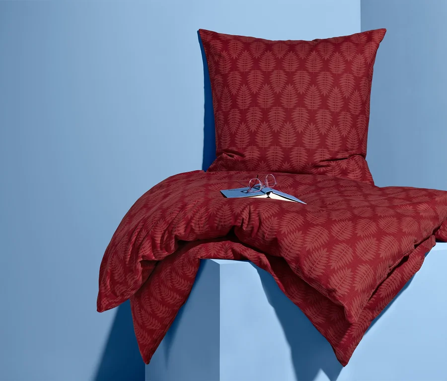 Ein Set Biber-Bettwäsche mit Blattmuster in Rot liegt auf einem blauen Sockel, darauf liegen ein Buch und eine Brille.