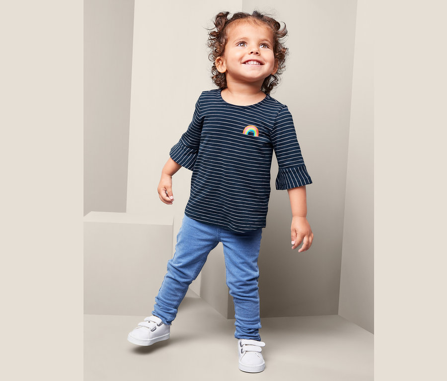 Ein Mädchen trägt ein Streifen-Shirt mit Regenbogen, blaue Jeans und weiße Schuhe.