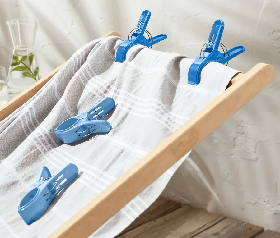 Eine gestreifte Decke ist mit vier blauen Handtuchclips an einer zusammenklappbaren Liege befestigt.