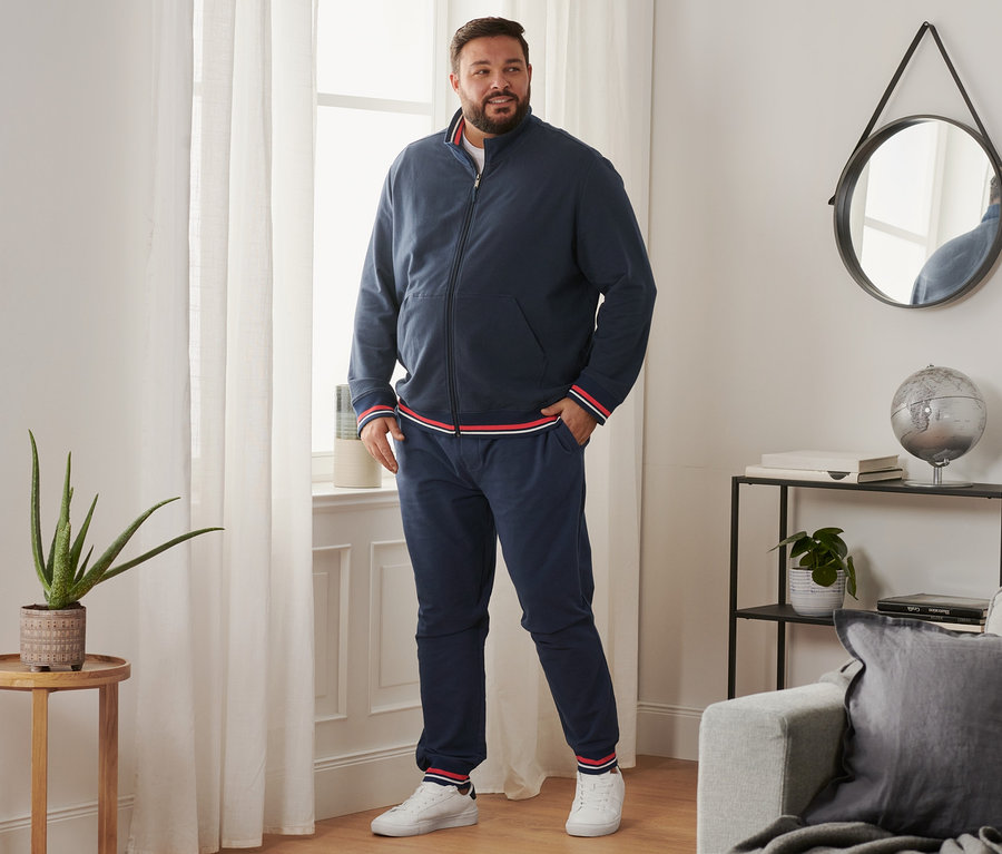 Mann steht am Fenster und trägt eine Sweathose, Sneaker und eine Sweatjacke.