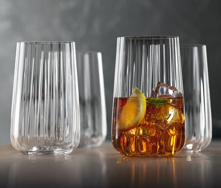 Vier Spiegelau Longdrinkgläser »Lifestyle« stehen auf einem Tisch, eines davon ist mit einem Getränk, Eis und einer Zitronenscheibe gefüllt.