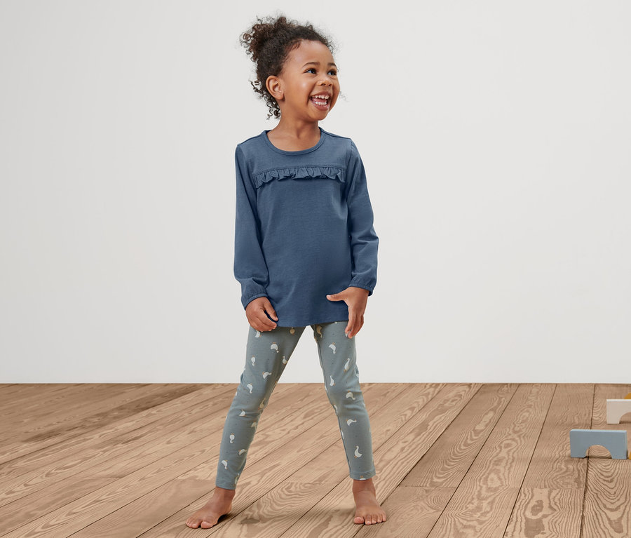Lächelndes kleines Mädchen steht auf einem Holzboden und trägt ein Kleinkind-Shirt und Leggings.