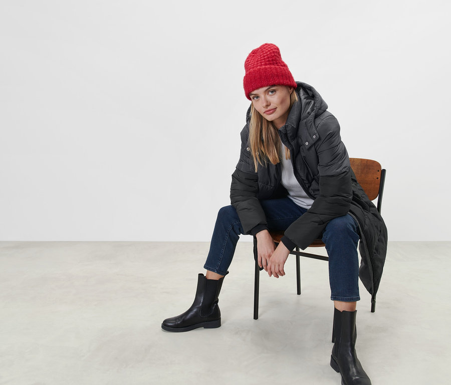 Frau sitzt auf einem Stuhl und trägt einen Steppmantel mit Kapuze und eine rote Mütze.