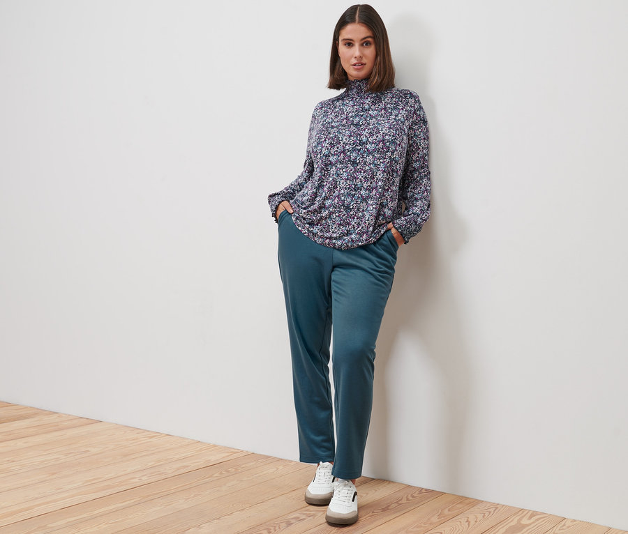 Frau posiert in Blusenshirt mit Stehkragen, millefleurs, Joggpants und Leder-Sneakern.