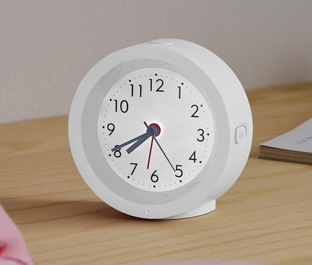 Ein weißer analoger Quarzwecker mit Hintergrundbeleuchtung steht auf einem Nachttisch neben einem Bett.