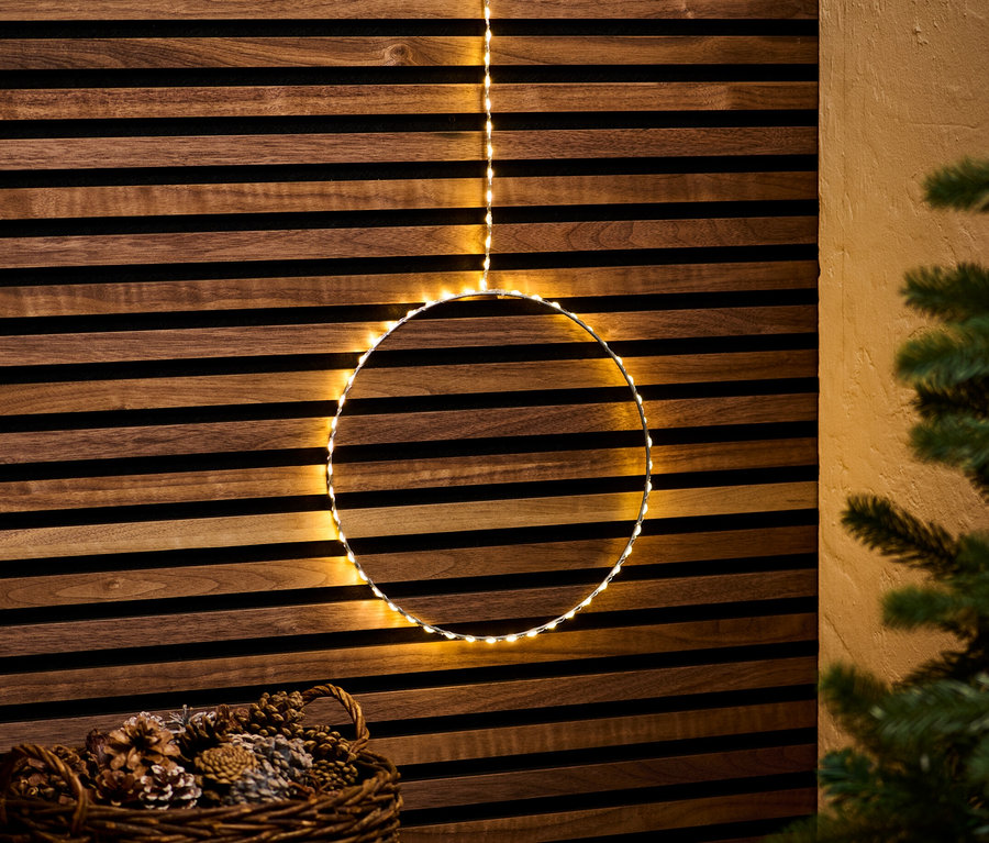 Ein hängender Outdoor-LED-Kreis vor einer Holzwand, darunter ein Korb mit Tannenzapfen.