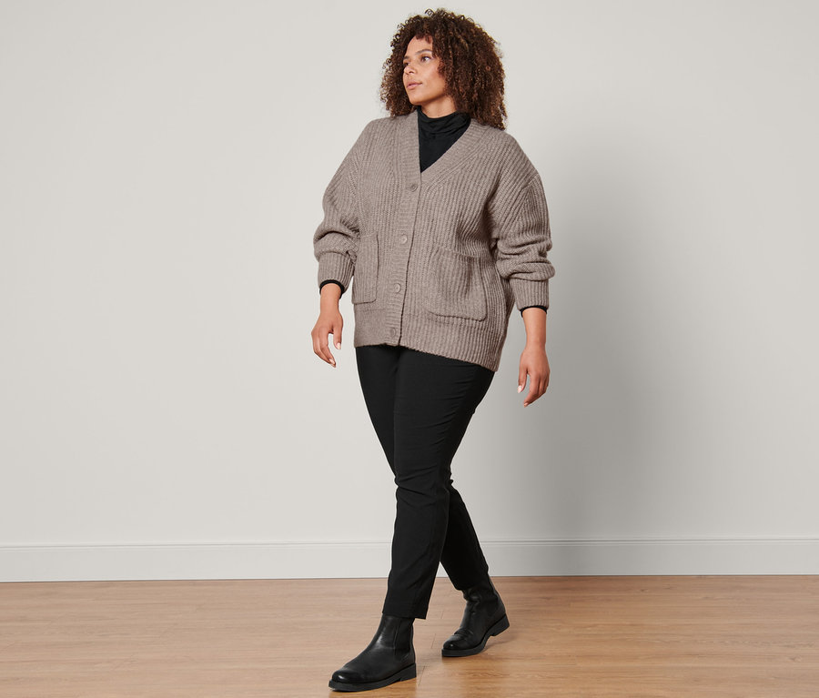 Eine Frau geht und trägt einen Grobstrick-Cardigan mit Wolle, ein schwarzes Rollkragenshirt, eine schwarze Konfektionierte Bengalinhose und Lederboots.