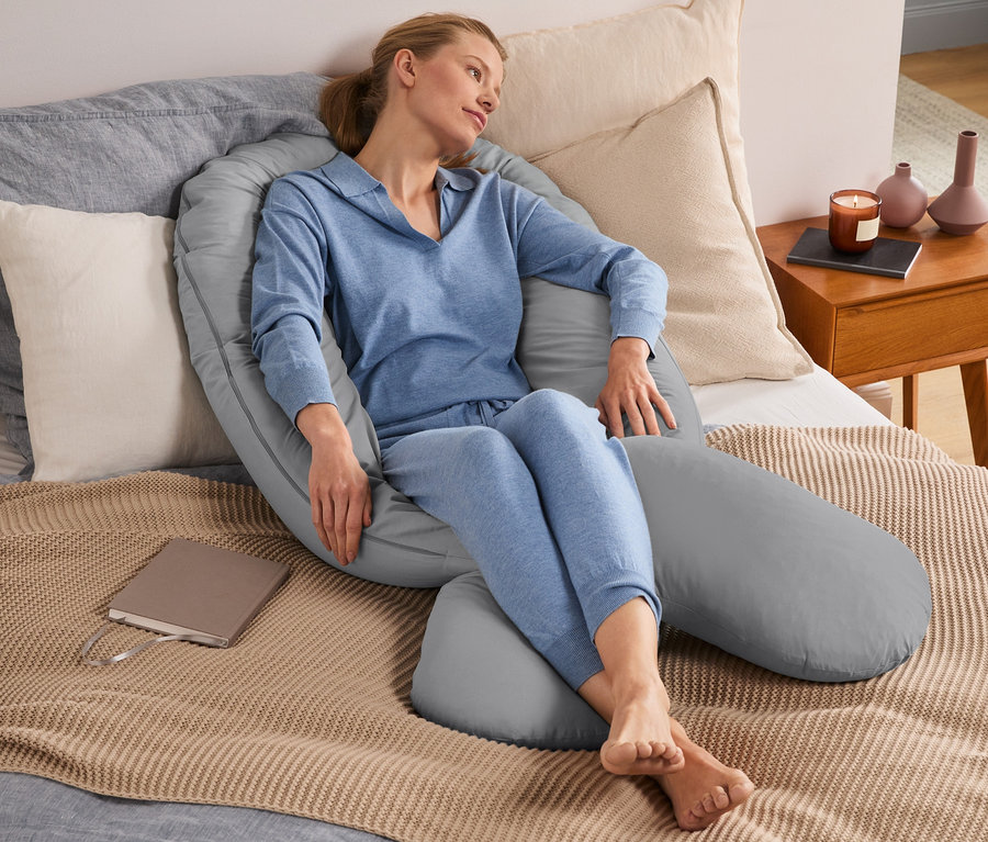 Eine Frau sitzt im Bett, umgeben von einem grauen Multifunktionskissen XXL.
