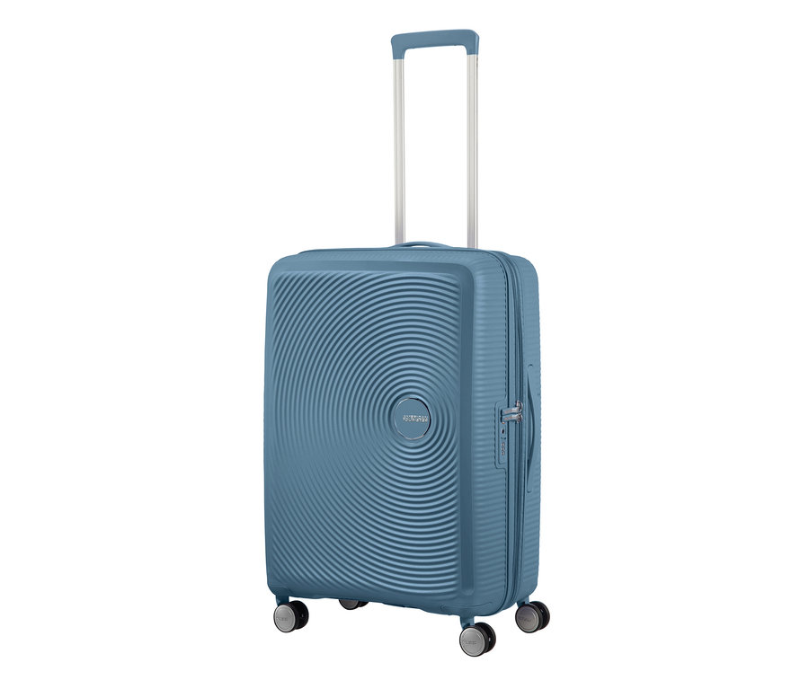 Ein mittelgroßer, blauer American Tourister Hartschalen-Koffer »Soundbox« Spinner mit silbernem Teleskopgriff und Rollen.