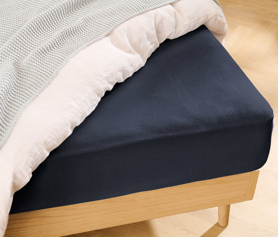 Detailansicht eines dunkelblauen Jersey-Spannbettlakens in Doppelgröße auf einem Bett mit weißer Bettdecke und grauer Decke.