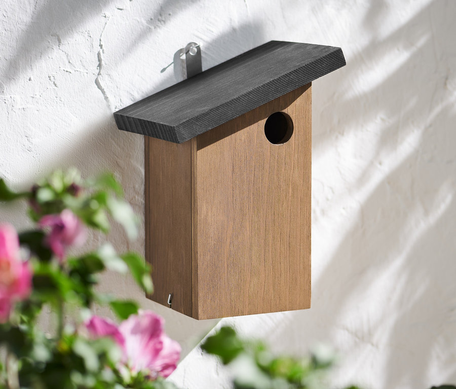 Ein modernes Vogelhaus hängt an einer weißen Wand, im Vordergrund rosa Blüten.