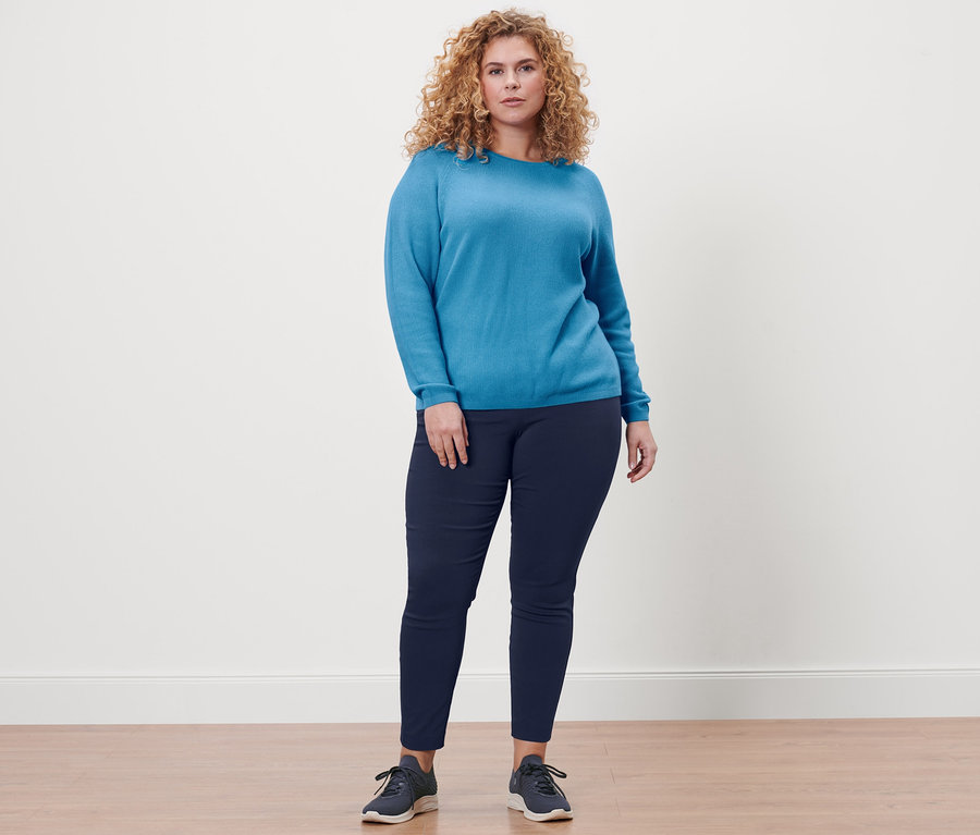 Frau steht und trägt eine dunkelblaue Stretchhose und einen azurblauen Strickpullover.