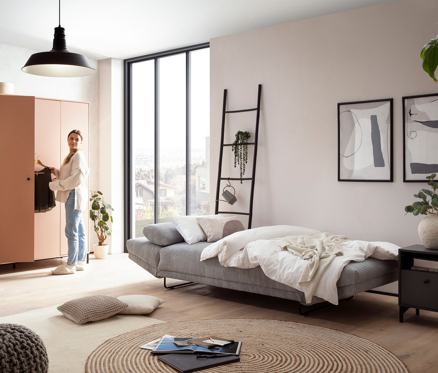 Frau greift in einem Wohnzimmer in einen Kleiderschrank, wo ein graues DOMO collection Daybed »Molde Leif55« steht.