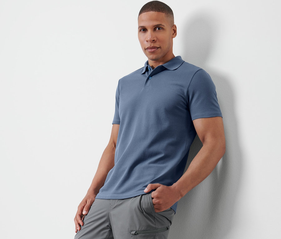 Mann lehnt an Wand, trägt Funktionspoloshirt und graue Hose.