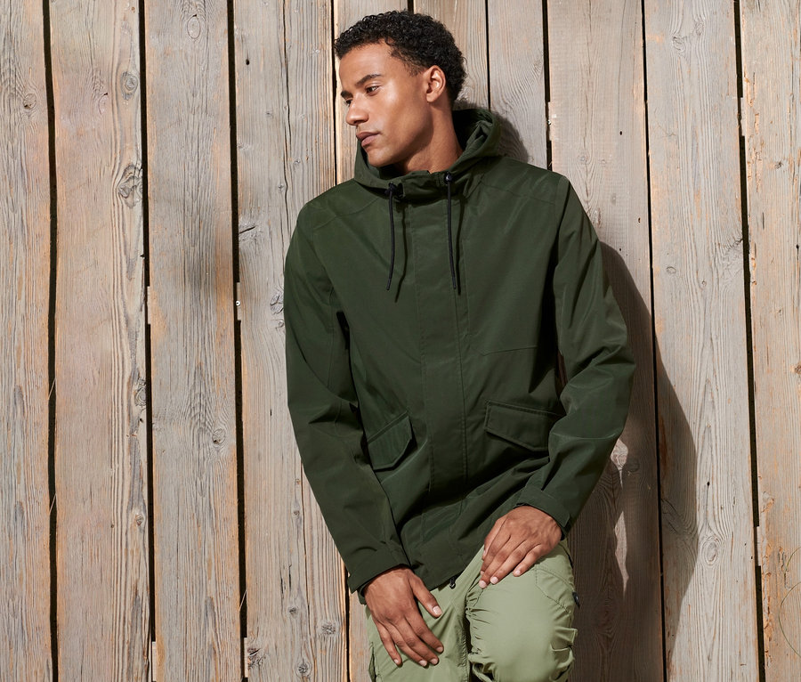 Mann lehnt an Holzwand und trägt einen Funktionsparka.