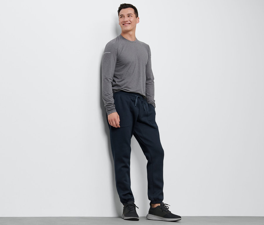 Mann lehnt in grauem Shirt und einer Sweathose an der Wand.