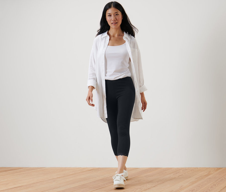 Frau geht und trägt eine schwarze 3/4-Leggings mit Spitzenborte, ein weißes Tanktop und ein weißes Hemd.