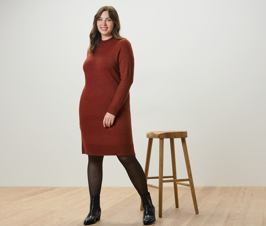 Frau posiert in einem Strickkleid mit Wollanteil und einer 50-DEN-Feinstrumpfhose in transparenter Optik neben einem Holzhocker.