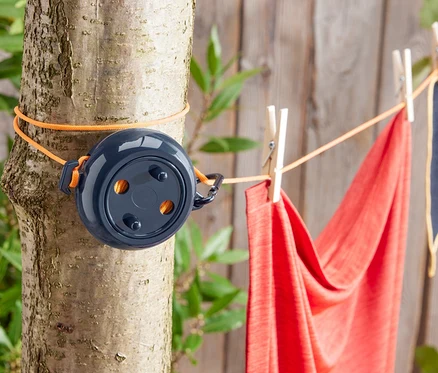 Eine Camping-Wäscheleine ist mit einer orangefarbenen Schnur an einem Baum befestigt. An der Leine hängen mit Wäscheklammern ein rotes und ein blaues Handtuch.