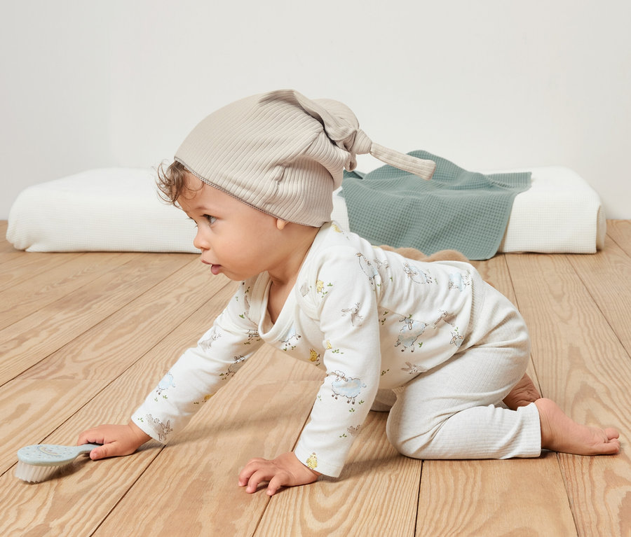 Baby krabbelt auf einem Holzboden und trägt eine beige Jerseymütze.
