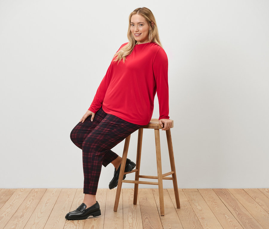 Frau sitzt auf einem Hocker und trägt eine karierte Stretchhose und ein Blusenshirt mit Raffung.