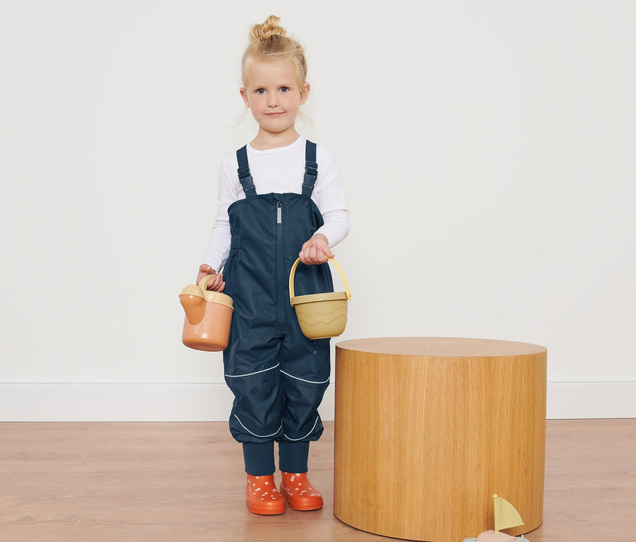 Ein Mädchen in Kleinkinder-Regenhose hält Spielzeug neben einem Holztisch.